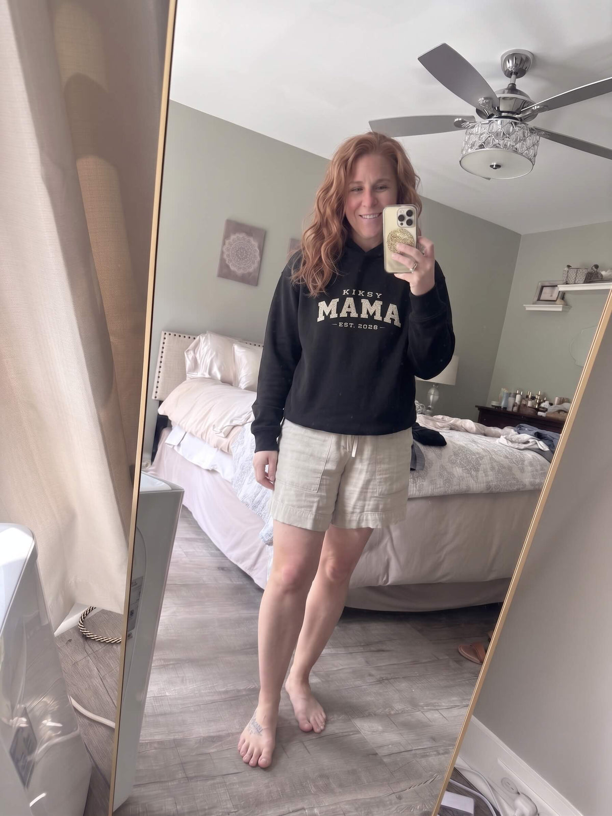 Woman wearing a black Kiksy Mama Est 2026 hoodie with distressed white varsity lettering.