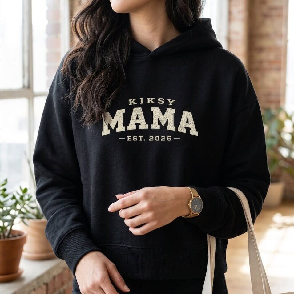 Kiksy black Mama Est 2026 hoodie worn by a woman indoors, featuring vintage varsity lettering and cozy fleece fabric.