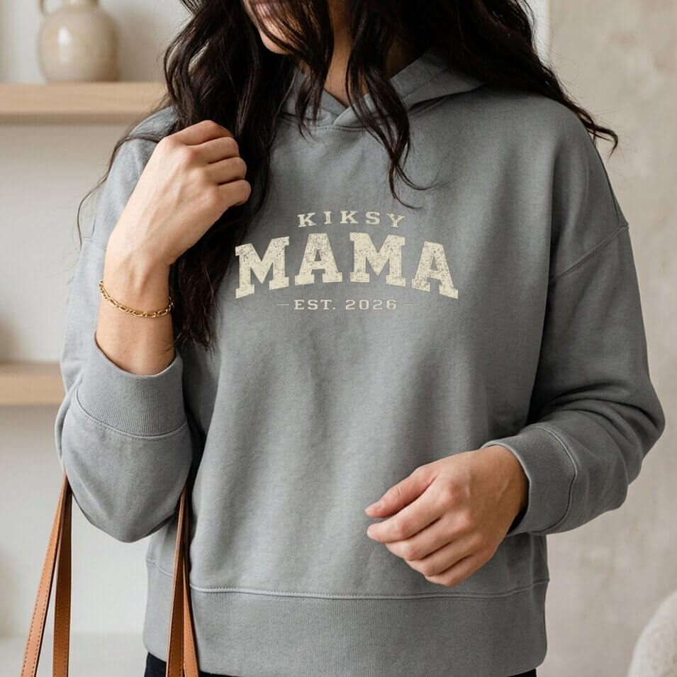 Kiksy grey Mama Est 2026 hoodie worn by a woman indoors, featuring vintage varsity lettering and cozy fleece fabric.