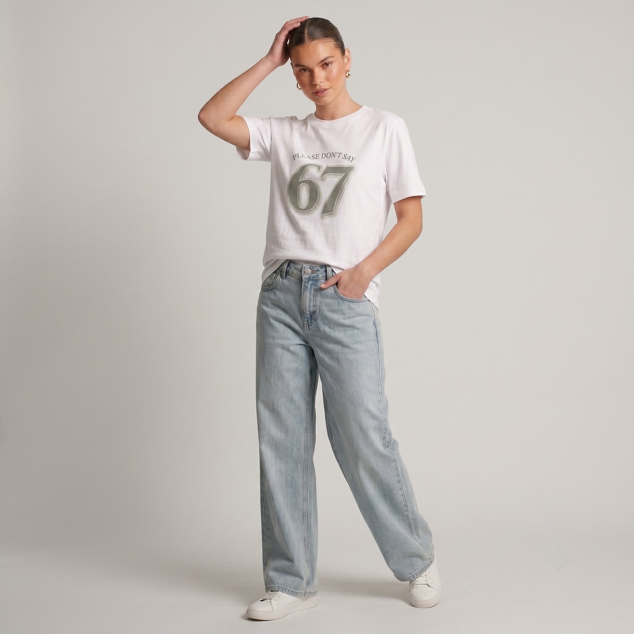 Woman wearing Kiksy white relaxed slogan t-shirt featuring Please Don't Say 67 print with casual blue jeans.